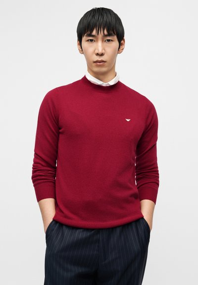 Κόκκινο πουλόβερ με στρογγυλή λαιμόκοψη, κατασκευασμένο από λεπτό πλεκτό ύφασμα με ριμπ μανσέτες και τελείωμα, με μικρό λογότυπο στο αριστερό στήθος. Συνδυάζεται με παντελόνια με ρίγες.