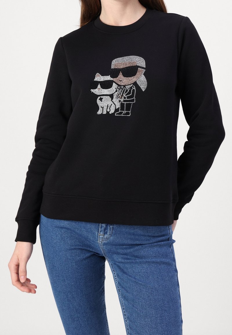 Sweat-shirt noir avec un motif en strass représentant une personne portant des lunettes de soleil et un chien également avec des lunettes de soleil, associé à un jean bleu.