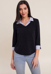 Haut noir à col en V avec des manches trois-quarts et des poignets à rayures bleu clair. Tissu texturé avec une finition lisse, associé à un denim bleu clair.