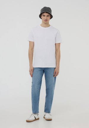 T-shirt bianca in cotone abbinata a jeans di denim azzurro chiaro. Un cappello a secchiello grigio completa l'outfit, accompagnato da sneaker bianche con dettagli beige.