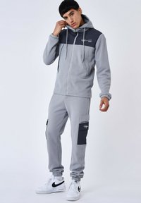 Tuta in pile grigio con un pannello contrastante grigio scuro, felpa con zip, polsini elasticizzati e tasche cargo; abbinato a sneakers bianche.