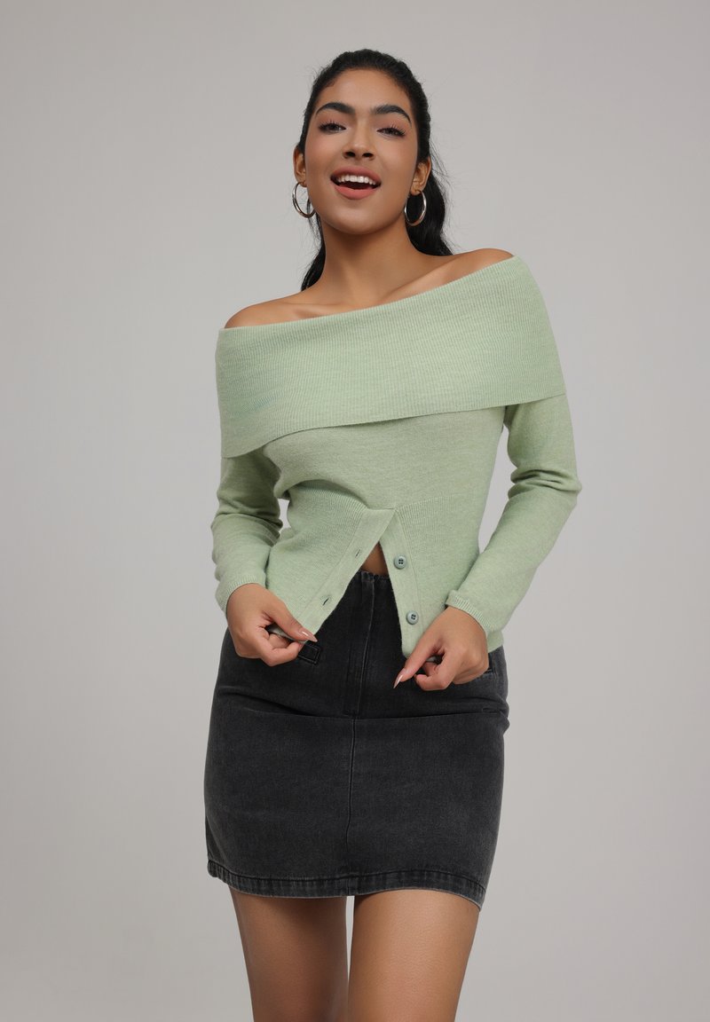 Green off-the-shoulder sweater with long sleeves, textured knit, and button closure. Worn with a high-waisted black denim skirt.
