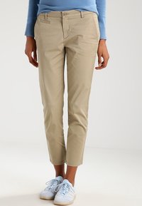 Person som bär beige slim-fit korta byxor, ljusblå tröja och vita sneakers med ljusblå snören, stående på ett vitt golv.