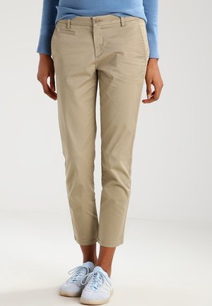 Person som bär beige slim-fit korta byxor, ljusblå tröja och vita sneakers med ljusblå snören, stående på ett vitt golv.