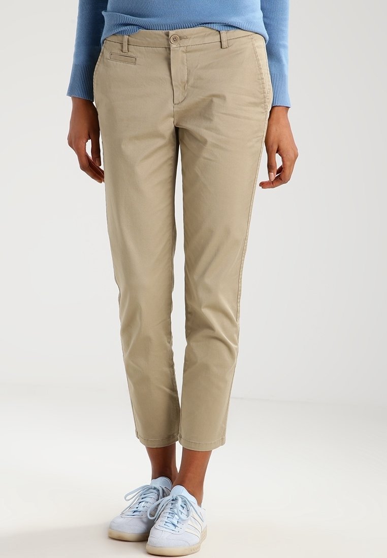 Person som bär beige slim-fit korta byxor, ljusblå tröja och vita sneakers med ljusblå snören, stående på ett vitt golv.