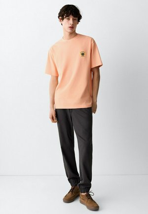 Junger Mann trägt ein pfirsichfarbenes T-Shirt mit kleinem Logo, dunkle Jogginghosen und braune Schnürschuhe, steht vor einem schlichten weißen Hintergrund.