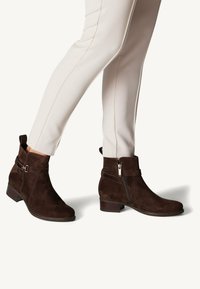 Bottines en daim marron avec une fermeture éclair sur le côté, des détails de sangle et un petit talon empilé, associées à un pantalon slim crème.