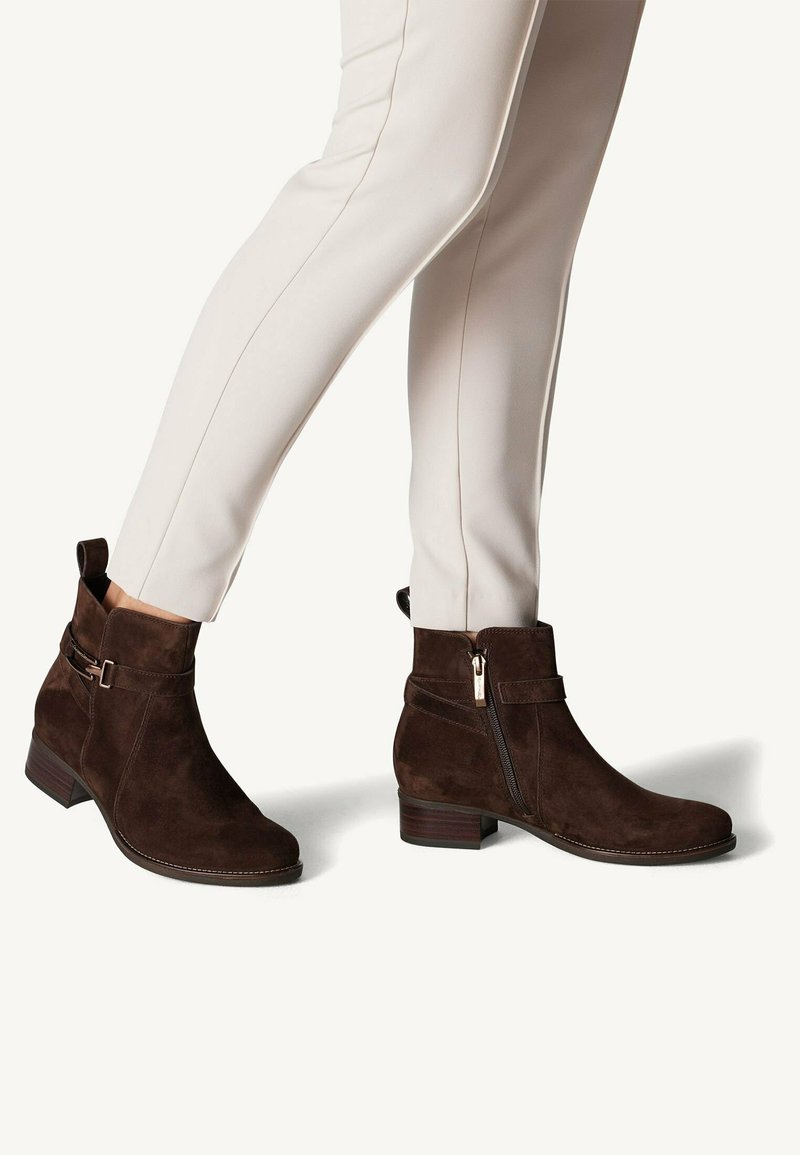 Bottines en daim marron avec une fermeture éclair sur le côté, des détails de sangle et un petit talon empilé, associées à un pantalon slim crème.