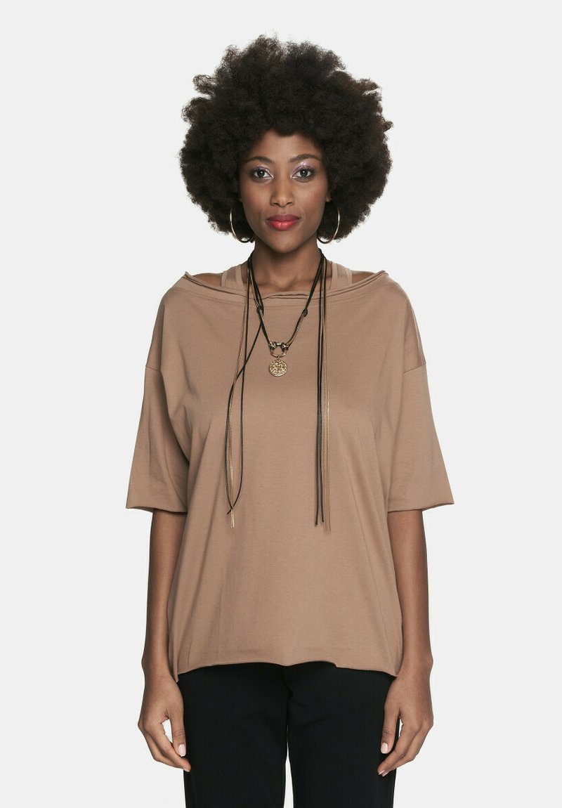 Brązowa, oversize krótka bluzka z okrągłym dekoltem i fakturowanym materiałem, zestawiona z długim naszyjnikiem z wisiorkiem i frędzlami.