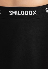 Mustat urheilushortsit, joissa on leveä vyötärönauha ja valkoinen "SMILODOX" -brändäys. Kangas on sileää ja joustavaa, suunniteltu mukavuutta varten.