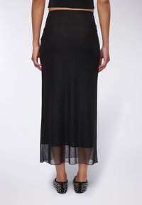 Gonna lunga nera in tessuto morbido e elasticizzato. Caratterizzata da un orlo inferiore trasparente e una cintura a scallop. Abbinata a scarpe nere con borchie.