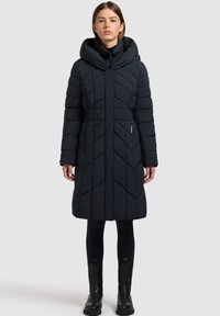 Vrouw draagt een lange zwarte doorgestikte donsjas met een hoge kraag, gecombineerd met zwarte legging en zwarte enkellaarsjes, staand tegen een effen achtergrond.