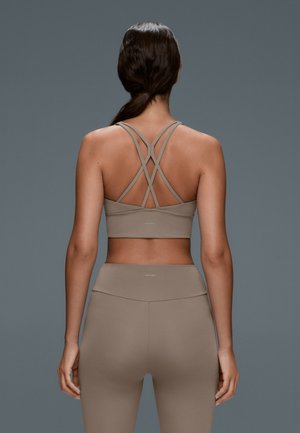 Femme aux cheveux foncés en queue de cheval basse, portant un brassière de sport beige à bretelles croisées et un legging taille haute assorti, debout devant un fond gris.