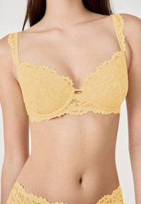 Reggiseno giallo in pizzo con motivi floreali, bordi scallop e spalline decorative, dal design strutturato e con supporto a ferretto.