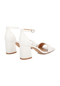 Sandali bianchi con tacco a blocco, cinturini alla caviglia e punta aperta. Materiale in pelle liscia, suola piatta marrone e design minimalista.