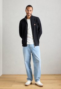Hombre con gafas de pie, vistiendo una chaqueta negra con cremallera, camiseta blanca, jeans azul claro y zapatillas blancas sobre un suelo de madera.