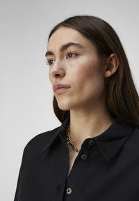 Camisa negra de botones con cuello y una tela texturada. Presenta botones negros y un collar de cadena plateada gruesa como acento.