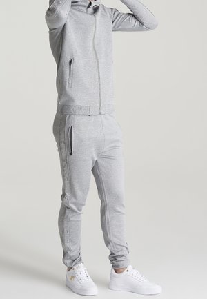 Survêtement gris clair en tissu doux et texturé, avec poches zippées, silhouette ajustée et détails à boutons-pression sur le pantalon.