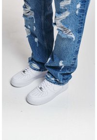 Weiße Ledersneaker mit einem perforierten Vorderfußbereich, kombiniert mit zerrissenen blauen Jeans aus Denim, die mehrere Risse und einen ausgefransten Saum aufweisen.