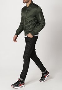 Veste bomber kaki en satin lisse, fermeture éclair à l'avant, poches latérales, portée avec un pantalon noir ajusté et des baskets noires et rouges.