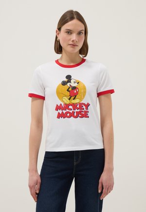 Mujer vistiendo camiseta blanca con ribete rojo con gráfico y texto de Mickey Mouse, combinada con vaqueros azul oscuro, posando contra un fondo liso.