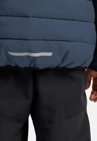 Marinblå pufferjacka med horisontella quiltade mönster, har en silverreflekterande remsa vid nederkanten. Tyget har en slät, väderbeständig yta.