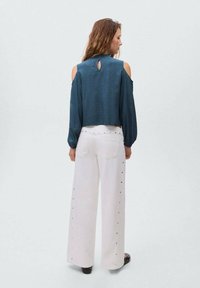 Bluză cu modele în nuanță de teal, cu umeri descoperiți și un decolteu în partea din spate, purtată împreună cu pantaloni largi alb cu talie înaltă, accentuați cu știfturi argintii.