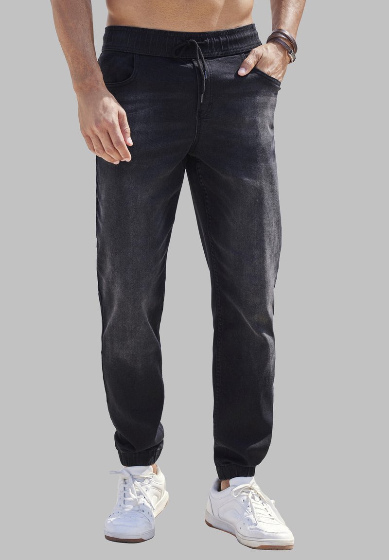 Pantalones jogger de mezclilla negra con cintura elástica, cierre de cordón, bolsillos laterales y tobillos ajustados, combinados con zapatillas blancas.