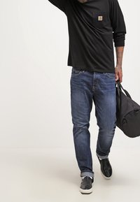 Schwarzes Langarmshirt mit Brusttasche, blaue Jeans und schwarze Slipper. Trägt eine schwarze Tasche und steht vor einem hellen Hintergrund.