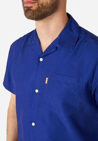 Camicia a manica corta con bottoni in cotone blu brillante, dotata di taschino sul petto, con bottoni bianchi e una piccola etichetta arancione sulla tasca.