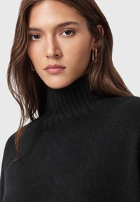 Suéter de cuello alto negro con neckline acanalado, hecho de una suave tela de punto. El modelo tiene el cabello largo y lleva pendientes de aro dorados.