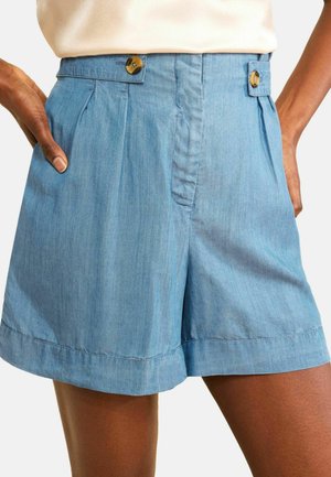 Pantaloni scurți bleu deschis cu talie înaltă, cu nervuri laterale cu nasturi, pliuri și buzunare laterale, purtați de o persoană cu mâinile parțial în buzunare.