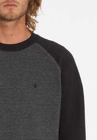 Sweat-shirt gris avec des manches raglan noires, présentant une texture subtile et un petit logo sur la poitrine. Encolure et poignets côtelés.