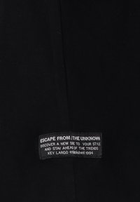 Czarna tkanina z naszytym prostokątnym metkiem z napisem "Ucieknij od nieznanego, odkryj nową stronę swojego stylu, wyprzedź trendy, Key Largo 1994."