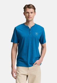 Modrá pruhovaná krátkásleeve henley košile vyrobená z lehkého materiálu, s výstřihem na tři knoflíčky a malým logem na levé straně hrudi.