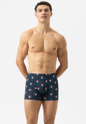 Muskulöser Mann in marineblauen Boxershorts mit rot-weißer Blumenmuster, der mit den Händen hinter dem Rücken vor einem schlichten weißen Hintergrund steht.