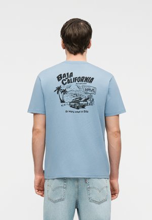 Noor inimene kannab helerohelist T-särki, millel on must Baja California rannamaastik, veoauto, palmipuud ja tekst seljal.