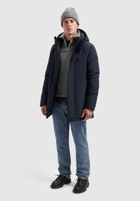 Marineblauwe pufferjack met een capuchon, een gestructureerde voorkant en zwarte accenten, gelaagd over een grijze gebreide trui, gecombineerd met blauwe jeans en zwarte schoenen.