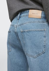 Vista posteriore di una persona che indossa jeans in denim azzurro con etichetta in pelle Massimo Dutti e top grigio scuro.