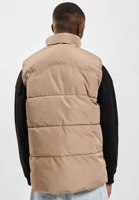 Beige puffers læg med højt krave, quiltet design og synlige sømme. Båret over en sort langærmet skjorte og lyse denim.