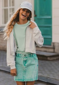 Grijze cropped hoodie, mintgroene t-shirt en lichtblauwe denimrok met vervaagde textuur. Blauwe pet met bloemembroderie. Lachende uitdrukking.