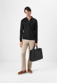 Zwart langemouwen polo met rits, beigen broek, bruine instappers en een zwarte leren boodschappentas met verschillende vakken.