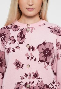 Rosa blomsterbelagt cardigan med rund hals, knappelukking og strukturet strikket stoff med mørkere burgunder aksenter.