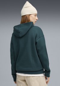 Felpa con cappuccio color turchese, dalla vestibilità comoda, con texture morbida e polsini a coste. Indossata sopra una maglietta bianca, abbinata a pantaloni beige chiaro.