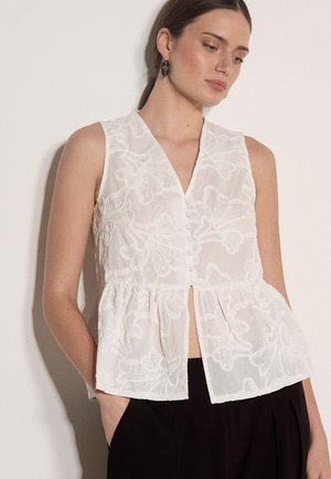 Frau trägt ärmellose weiße bestickte Bluse mit Knöpfen und schwarze Hose, steht vor einem schlichten hellen Hintergrund.