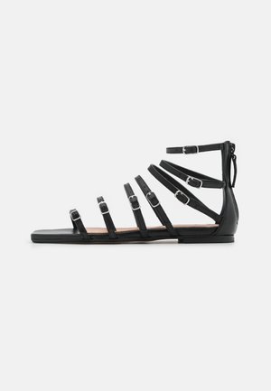 Sandale gladiateur plate noire avec six fines brides réglables et boucles argentées, fermeture éclair arrière et design à bout ouvert.