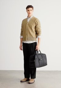 Brūns vilnas kardigans, melna soma ar dubultiem rokturiem, svītrainais krekls, melnas bikses, bēšas kurpes, stāv uz gludas gaišas grīdas.