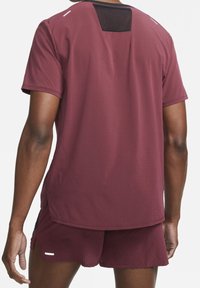 Camiseta deportiva burdeos con un diseño texturizado, mangas cortas y un panel de malla negra en la parte trasera. Incluye aberturas laterales para mayor transpirabilidad.