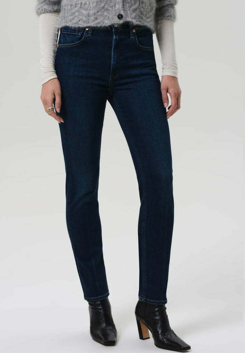 Jean en denim bleu foncé à coupe slim, avec des détails de couture subtils et une taille haute, associé à des bottines noires à talons.