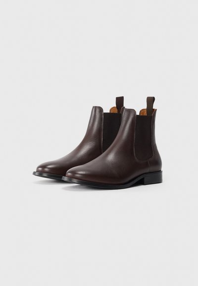 Un par de botas Chelsea de cuero marrón oscuro con paneles laterales de elastano negros y pestañas para tirar, sobre un fondo blanco.